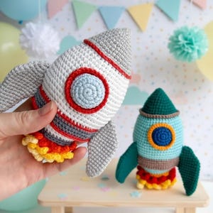 May include: Two handmade crochet rockets. One is white with red, gray, and blue accents, and the other is teal with green, brown, and yellow accents. Both have wings and flame details, perfect for a child's toy or nursery decor.