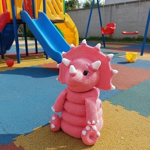 Op de afbeelding: Een roze gehaakte Triceratops dinosaurus pluche speelgoed met witte hoorns en accenten. Het handgemaakte knuffeldier zit op een kleurrijk speeloppervlak. Het speelgoed is een schattig en knuffelbaar item.