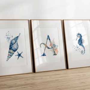 Könnte beinhalten: Drei gerahmte Aquarelldrucke mit einer Muschel, dem Buchstaben A und einem Seepferdchen. Die Drucke sind in Blautönen und Orange.