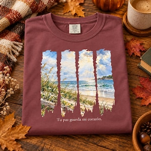 May include: A maroon crewneck t-shirt featuring a graphic of a coastal beach scene with tall grass, ocean waves, and a cloudy sky. The graphic is divided into four vertical strips. The text "Tu paz guarda mi corazón" is printed below.