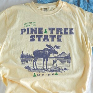 Könnte beinhalten: Hellgelbes T-Shirt mit dem Text "GREETINGS FROM THE PINE TREE STATE MAINE" und einer Elch-Grafik. Eine blaue Jeans liegt neben dem T-Shirt.