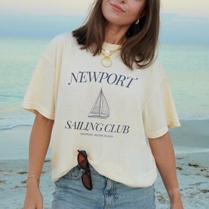 Peut inclure: Une femme portant un t-shirt crème avec le texte "NEWPORT SAILING CLUB NEWPORT, RHODE ISLAND" et un short en jean bleu. Elle est debout sur une plage de sable.