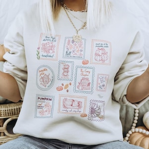 Könnte beinhalten: Ein cremefarbenes Sweatshirt mit Rundhalsausschnitt und einem Grafikdruck aus mehreren rechteckigen Briefmarkenmotiven. Jede Briefmarke enthält herbstliche Illustrationen und Texte wie "Apples", "Fall", "Pumpkin Patch", "Pumpkin Pie" und "Cozy Days" in Orange-, Blau- und Rosatönen.