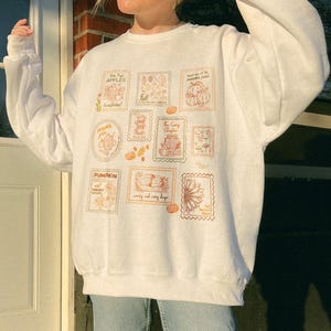 Puede incluir: Sudadera blanca con un diseño de sellos con temática otoñal que presenta calabazas, manzanas y el texto "Pumpkin". La sudadera se combina con vaqueros azul claro. El tema general es el otoño y la temporada.