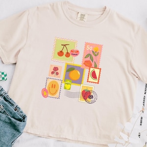 May include: Cream-colored t-shirt featuring a colorful stamp design with cherries, flowers, a peach, a watermelon slice, and a strawberry. The shirt is paired with blue jeans and a green and white checkered hair clip.