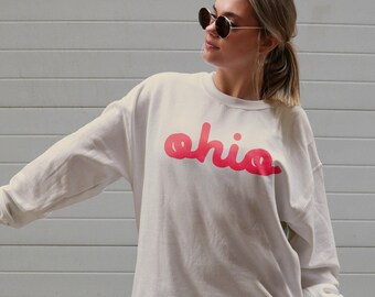 May include: A white sweatshirt with a red "ohio" graphic printed on the front. The sweatshirt is being worn by a person with long blonde hair and sunglasses.