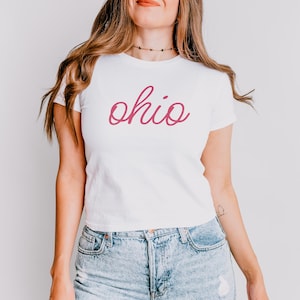 Puede incluir: Una camiseta blanca recortada con un gráfico de "Ohio" en cursiva rosa.