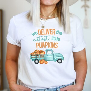 Op de afbeelding: Wit T-shirt met de tekst "we DELIVER the cutest little PUMPKINS" in teal en oranje. De afbeelding toont een lichtblauwe vrachtwagen vol met oranje pompoenen, met extra pompoenen voor de vrachtwagen.
