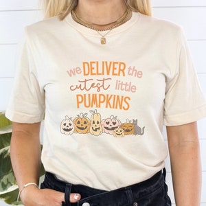 Puede incluir: Camiseta color crema con el texto "we DELIVER the cutest little PUMPKINS" en naranja y rosa. Debajo del texto hay calabazas de dibujos animados y un gato gris. La camiseta es de manga corta y cuello redondo.