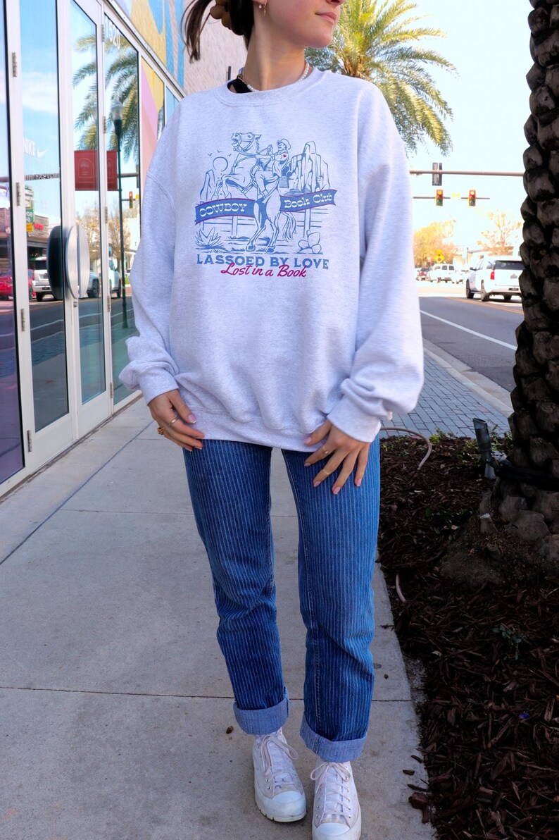 Cowboy Romance Book Club Graphic sweatshirt featuring a cowboy on a horse in a desert scene. the text reads Cowboy Book Club lassoed by love lost in a book.