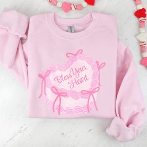 May include: A pink sweatshirt with a pink heart design and the text "Bless Your Heart" on the front.
