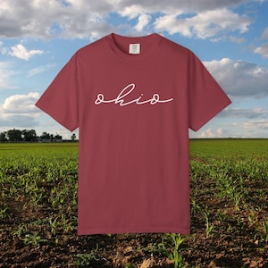 Puede incluir: Una camiseta granate con la palabra "ohio" en blanco en cursiva. La camiseta se muestra sobre un fondo de campo y cielo azul con nubes. La camiseta es de manga corta y cuello redondo.