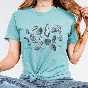 May include: A teal t-shirt featuring a collection of detailed seashell illustrations in black ink, including a crab and a seagull. The shirt has the phrases "Salt life" and "I never needed anything more" printed on it.