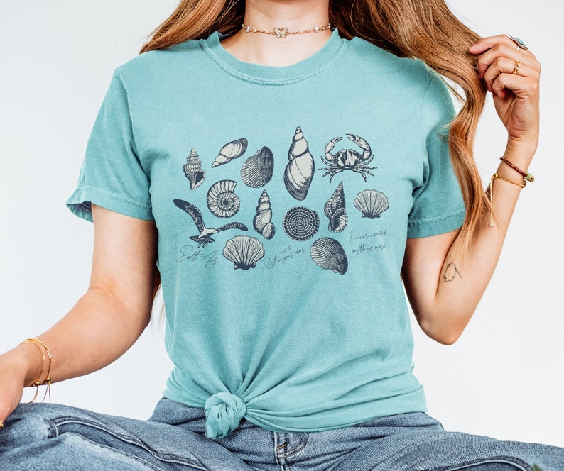May include: A teal t-shirt featuring a collection of detailed seashell illustrations in black ink, including a crab and a seagull. The shirt has the phrases "Salt life" and "I never needed anything more" printed on it.