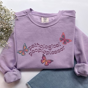 May include: A lavender sweatshirt with the text "pathological peoplepleaser" in a retro font. Three butterfly graphics are printed on the sweatshirt.