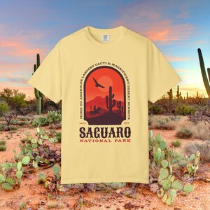以下が含まれることがあります： ペールイエローの半袖Tシャツ。サワロサボテン、鳥、馬に乗った人物が描かれた夕暮れの砂漠の風景グラフィックが特徴です。テキストには「HOME TO AMERICA'S LARGEST CACTI & MAGNIFICENT DESERT SUNSETS, ESTD 1994, SAGUARO NATIONAL PARK」と記されています。