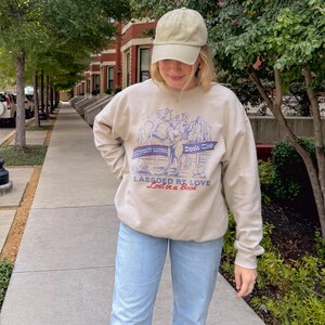 May include: Beige sweatshirt with the text "Cowboy Lovin' Book Club" and "Lassoed by Love Lost in a Book" in blue and red. The sweatshirt features a graphic of a cowboy roping a horse. The person is wearing a beige cap and light blue jeans.