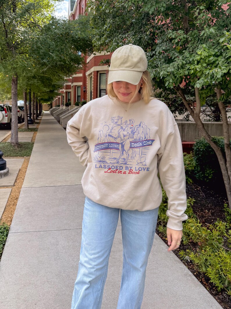 May include: Beige sweatshirt with the text "Cowboy Lovin' Book Club" and "Lassoed by Love Lost in a Book" in blue and red. The sweatshirt features a graphic of a cowboy roping a horse. The person is wearing a beige cap and light blue jeans.
