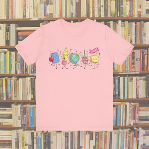 Könnte beinhalten: Ein hellrosa T-Shirt mit einem Grafikdruck von Cartoon-Schulmaterialien, darunter ein blaues Buch mit einem Apfel, ein gelber Textmarker, ein Globus, ein Eiskaffee und ein gelber Haftnotizzettel, der eine rosa Flagge mit dem Text TEACHER hält.
