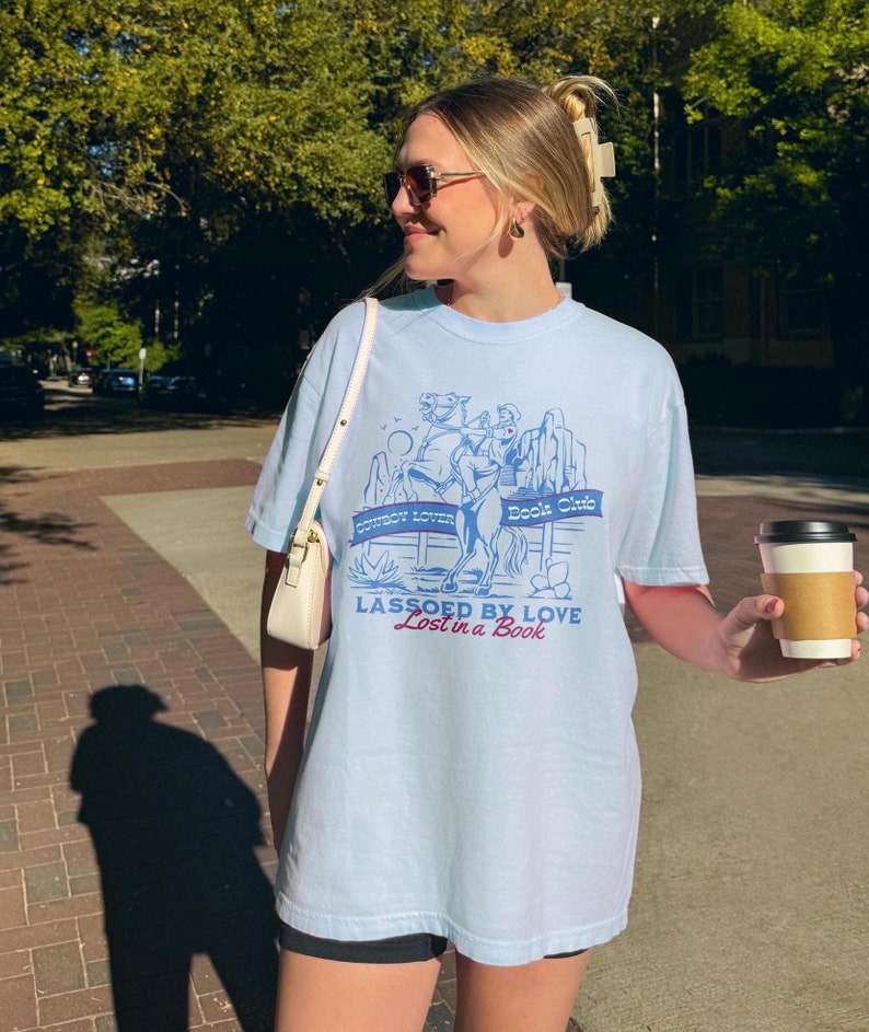 Pu&ograve; includere: Una maglietta blu chiaro con una grafica di un cowboy a cavallo. La grafica include il testo "Cowboy Lover Book Club" e "Lassoed by Love Lost in a Book".