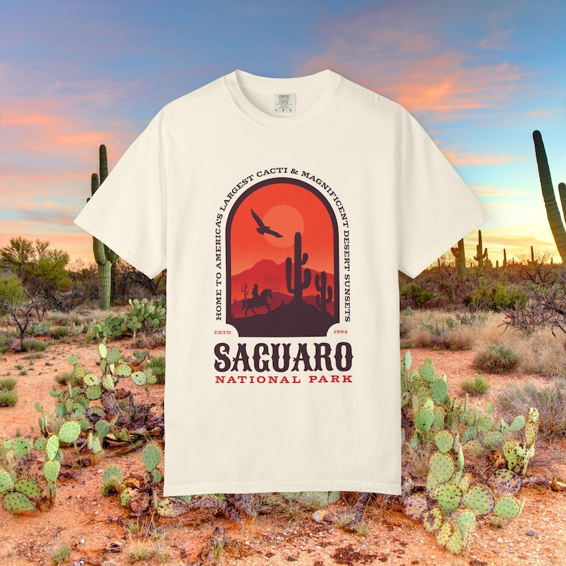 以下が含まれることがあります： オフホワイトの半袖Tシャツ。アーチの中に砂漠の風景が描かれています。デザインには、馬に乗った人のシルエット、サワロサボテン、鳥、そして「HOME TO AMERICA'S LARGEST CACTI & MAGNIFICENT DESERT SUNSETS」、「ESTD 1994 SAGUARO NATIONAL PARK」という文字が含まれています。