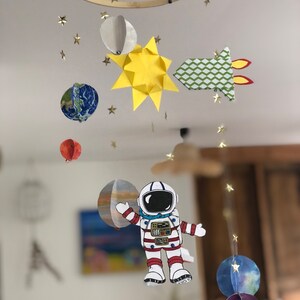 May include: A space-themed mobile with an astronaut, planets, sun, and rocket. The mobile has gold star accents and hangs from a wooden hoop. The planets are blue, orange, and multi-coloured. The rocket has a green and white chevron pattern.