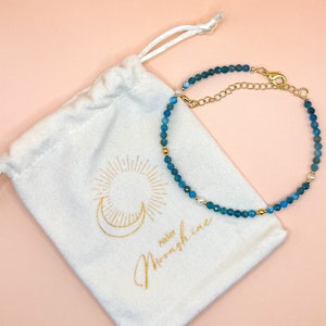 May include: A teal beaded bracelet with gold-coloured accents and a white drawstring bag. The bag has gold text reading "Atelier Moonshine" and a sun and moon design. The background is a light pink. The bracelet is made of small beads.