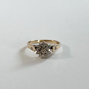 Könnte beinhalten: Ein goldener Ring mit einer zentralen Anordnung kleiner, runder, klarer Steine in einem Blumenmuster. Der Ring besteht aus poliertem Gold und enthält auf jeder Seite des Schafts in der Nähe des Clusters einen kleinen, quadratisch geschliffenen Stein.