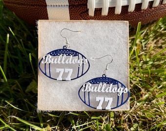 May include: A pair of dangle earrings shaped like American footballs, featuring a dark blue outline and white lettering that reads Bulldogs 77. The earrings are suspended from silver-toned metal hooks and are displayed on a square white card.