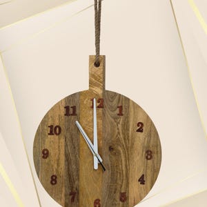 Op de afbeelding: Een ronde houten wandklok met een natuurlijke houtnerf afwerking. De klok heeft bruine cijfers en witte wijzers. Hij is opgehangen aan een bruin touw. De klok heeft een diameter van ongeveer 30 cm.