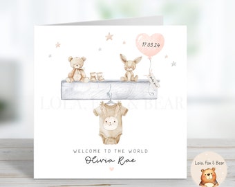 Puede incluir: Una tarjeta blanca con una ilustración de acuarela de un oso de peluche, un conejito y un body de bebé colgando de un estante. La tarjeta dice "Bienvenido al mundo Olivia Rae" y "17.03.24" con un globo en forma de corazón rosa. Una pequeña pegatina con un oso marrón y el texto "Lola, Fox & Bear" está en la esquina inferior derecha.