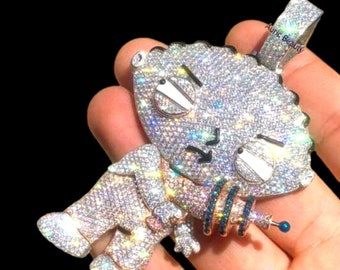 May include: A silver and white diamond-encrusted pendant featuring a cartoon character holding a gun. The pendant is on a silver chain.