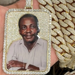 May include: A gold chain necklace with a large, square pendant. The pendant is covered in small diamonds and features a photo of a man inside. The chain is also covered in diamonds.