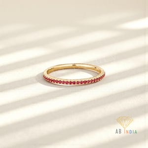 May include: A gold band ring with a continuous row of small, vibrant red gemstones. The ring is set against a soft, off-white background with subtle shadow patterns. The ring's design is simple and elegant, showcasing the gemstones.