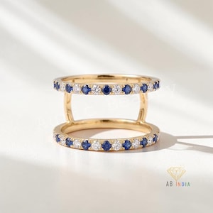 Puede incluir: Un anillo de oro de doble banda con dos bandas paralelas conectadas por barras verticales de oro. Cada banda está engastada con un patrón alterno de zafiros azules redondos y diamantes blancos transparentes. El texto Auric Beauty y AB INDIA es visible.