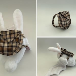 May include: A white plush bunny wearing a brown and tan plaid backpack. The backpack has a curved handle and a flap closure. The bunny's ears are upright, and the backpack is secured with straps.