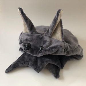 Könnte beinhalten: Eine graue Plüschfledermaus mit großen Ohren und schwarzen Augen. Die Fledermaus ist in einer Schlafposition zusammengerollt.