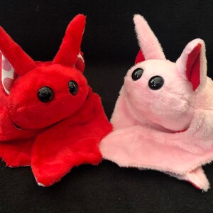 May include: Two plush bat toys, one red with white polka dots and one pink. Both have large black eyes and are lying on a black surface.