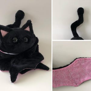 Puede incluir: Murciélago de peluche negro con alas de tela rosa y una cabeza de gato negra con grandes ojos negros. La cabeza del gato lleva un collar rosa.