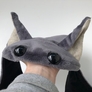Könnte beinhalten: Grauer Plüschhut mit großen schwarzen Augen und langen, schlaffen Ohren. Der Hut ist so gestaltet, dass er wie eine Fledermaus aussieht.