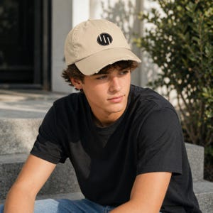 May include: A tan baseball cap with a distressed brim and a black circular logo featuring three vertical lines. The cap is made of a soft material and has a classic six-panel design. A casual accessory for everyday wear.