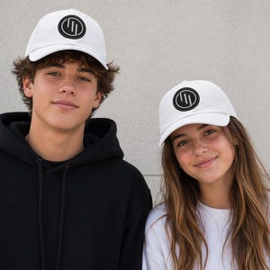 Pode incluir: Duas pessoas usando bonés de beisebol brancos com um logotipo circular preto. Os bonés têm uma aba curva e são feitos de tecido claro. Uma pessoa usa um moletom preto com capuz, a outra uma camisa branca.
