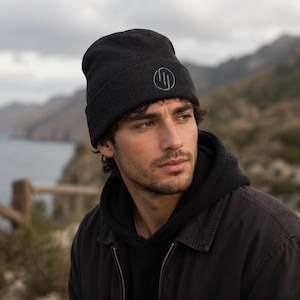 May include: A dark grey beanie hat with a circular emblem on the front. The beanie is made of a knitted fabric. The background shows a blurred landscape with mountains and a body of water.