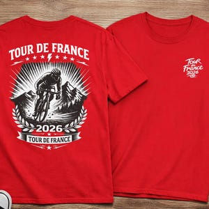 May include: Red t-shirts with a Tour de France theme. One shirt features a large graphic of a cyclist, mountains, and the text "Tour de France 2026." The other shirt has a smaller logo on the chest.