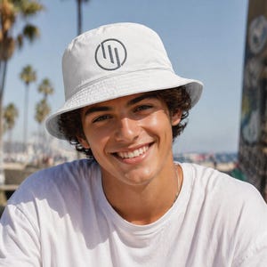 May include: White bucket hat with a dark grey circular logo. The hat is worn by a person wearing a white t-shirt. The background includes palm trees and a beach.