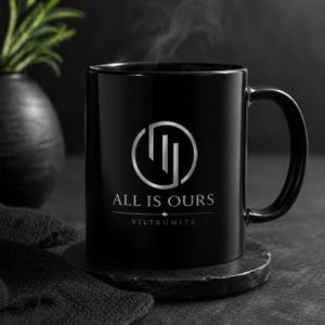 May include: A black ceramic mug with a silver logo and the text "ALL IS OURS" and "VILTRUMITE". The mug is sitting on a dark gray coaster, with steam rising from the top. A dark vase with a green plant is in the background.