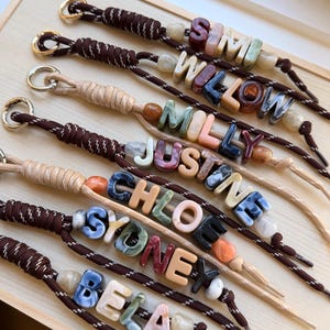 May include: A collection of seven wrist lanyards made from brown or beige cord, each featuring a gold-toned metal ring and personalised with colourful, marbled acrylic letter beads spelling the names SIMI, WILLOW, MILLY, JUSTINE, CHLOE, SYDNEY, and BELA.