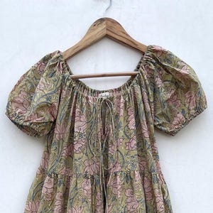 May include: A long, flowing dress with a floral pattern in shades of pink, green, and blue. The dress has short, puffed sleeves and a drawstring neckline. It is hanging on a wooden hanger.