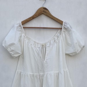 May include: A white tiered dress with short, puffed sleeves and a square neckline. The dress hangs on a wooden hanger against a plain white background. A drawstring detail is visible at the neckline.