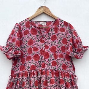 May include: A pink, tiered dress with a red floral pattern. The dress has short sleeves with ruffled edges and a mandarin collar. The fabric appears lightweight, suitable for warm weather. The dress is hanging on a wooden hanger.